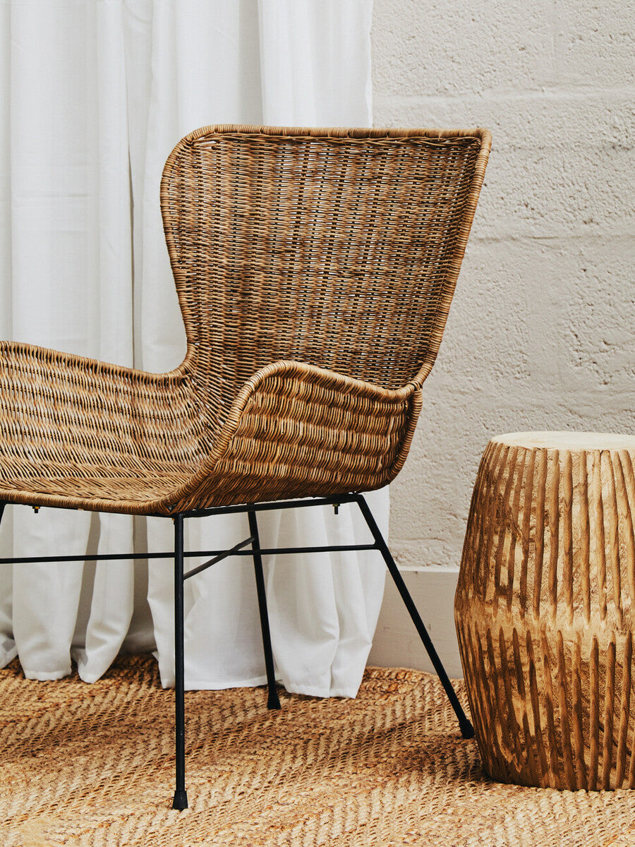Curved design natural rattan chair with a woven backrest and armrests, set on black metal legs, next to a wooden side table, against a light-colored wall and white curtains, with a wooden floor