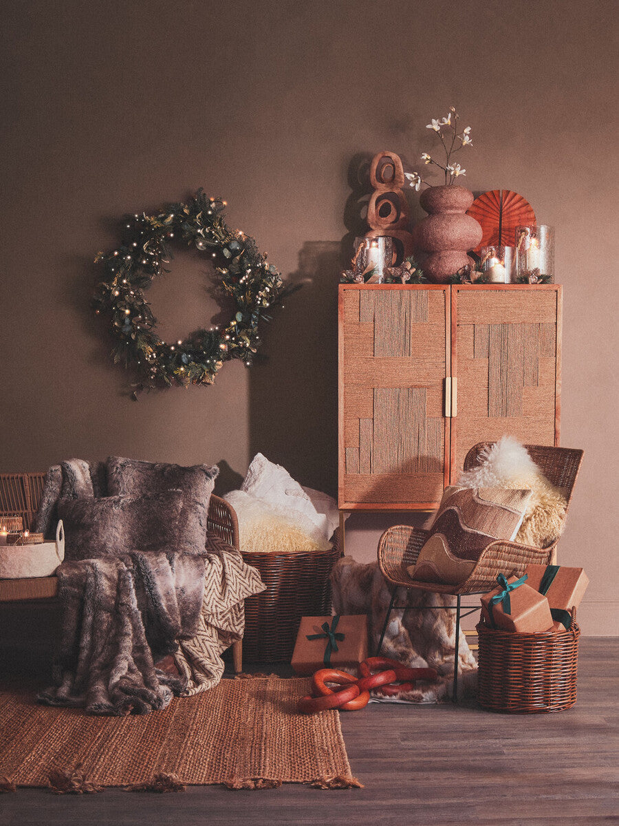 Curved design natural rattan chair positioned in a cozy living space, surrounded by decorative pillows, a wooden cabinet, and various textured items. A wreath hangs on the wall, adding a touch of greenery to the warm, inviting atmosphere