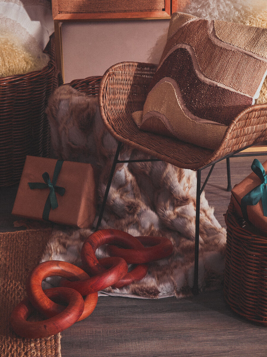 Curved design natural rattan chair with a light brown finish, surrounded by decorative hats, a red chain, and various textured materials, set against a warm, softly lit background
