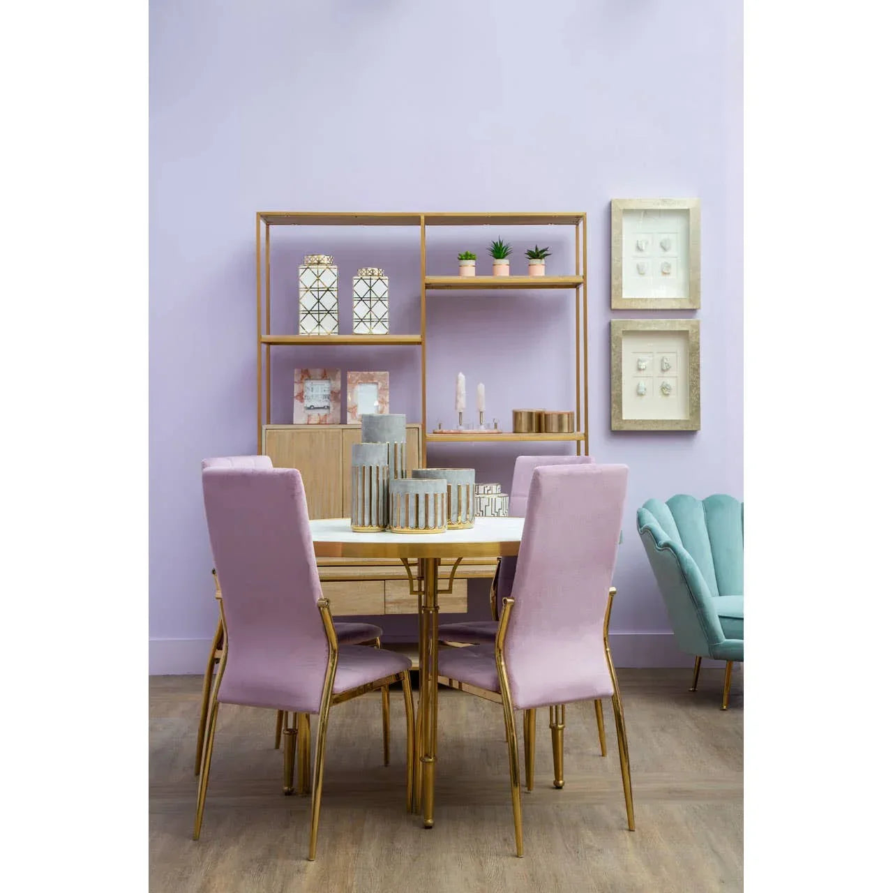 Modern dining table with gold legs, pink velvet chairs, and shelving against a lilac wall