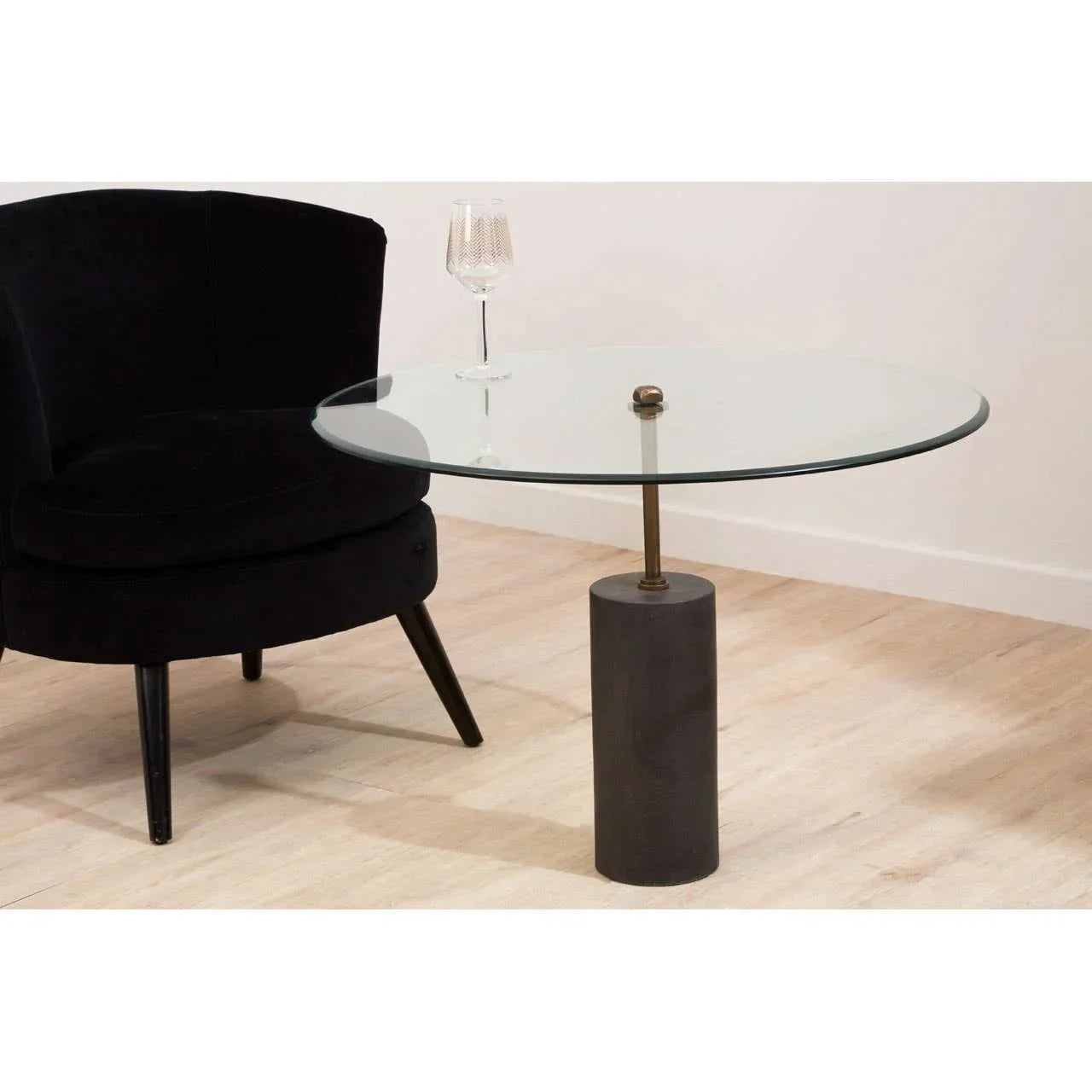 Round glass coffee table with cylindrical base beside black velvet chair on wood floor