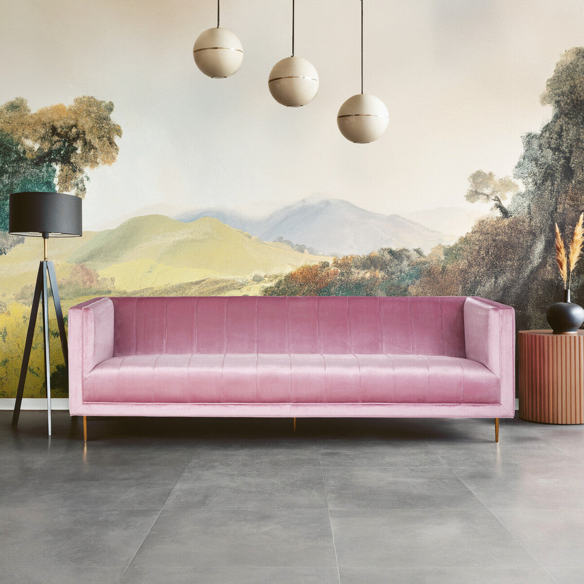 A three-seat pink sofa with a sleek, modern design featuring a smooth fabric upholstery and minimalist wooden legs. The sofa is positioned against a mural depicting rolling hills and trees, with three round pendant lights hanging above. A tall plant in a pot is visible to the right, complementing the contemporary decor