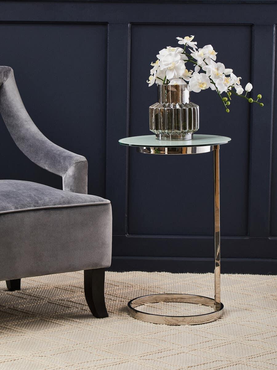 Round glass side table with chrome base beside grey velvet armchair on textured rug