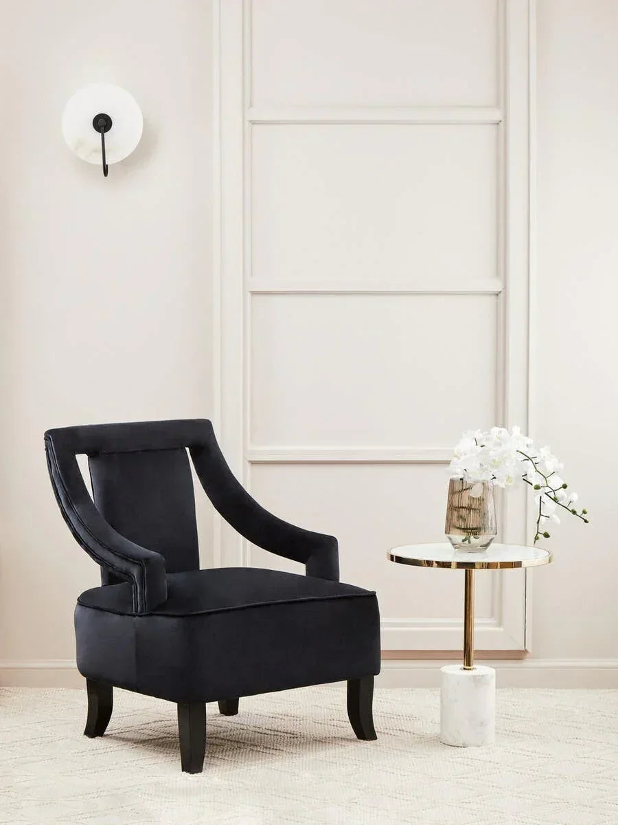 Black velvet accent chair with curved arms beside round gold side table and white orchid vase
