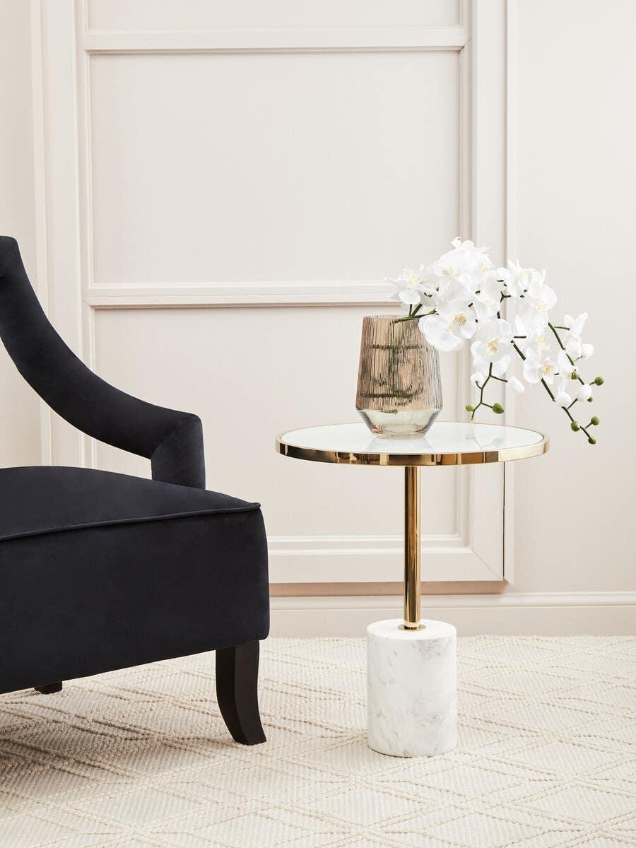 Round glass side table with gold metal frame and marble base beside black upholstered chair
