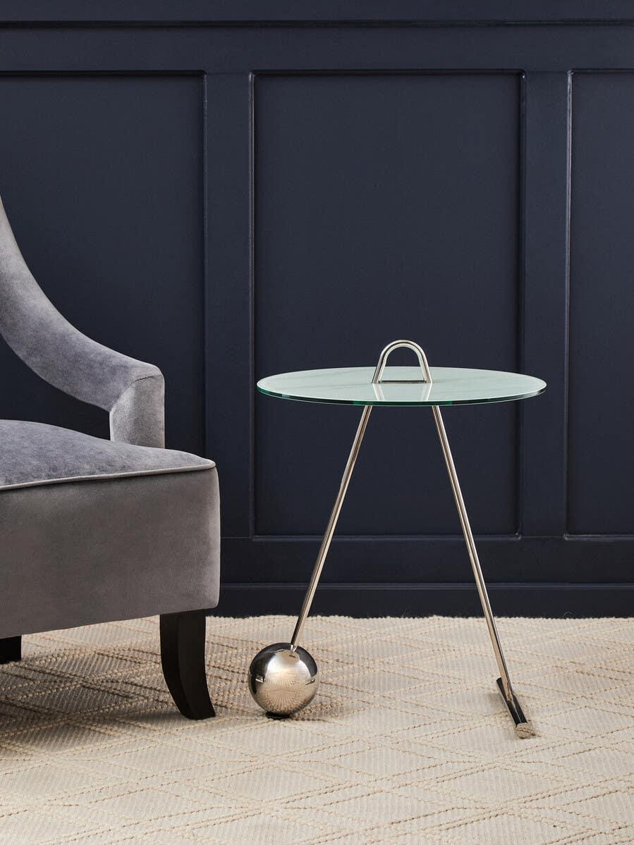 Modern glass side table with chrome legs beside a grey velvet chair on beige rug