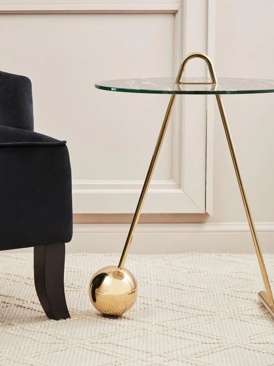 Modern glass top side table with gold metal frame beside navy velvet sofa on textured rug