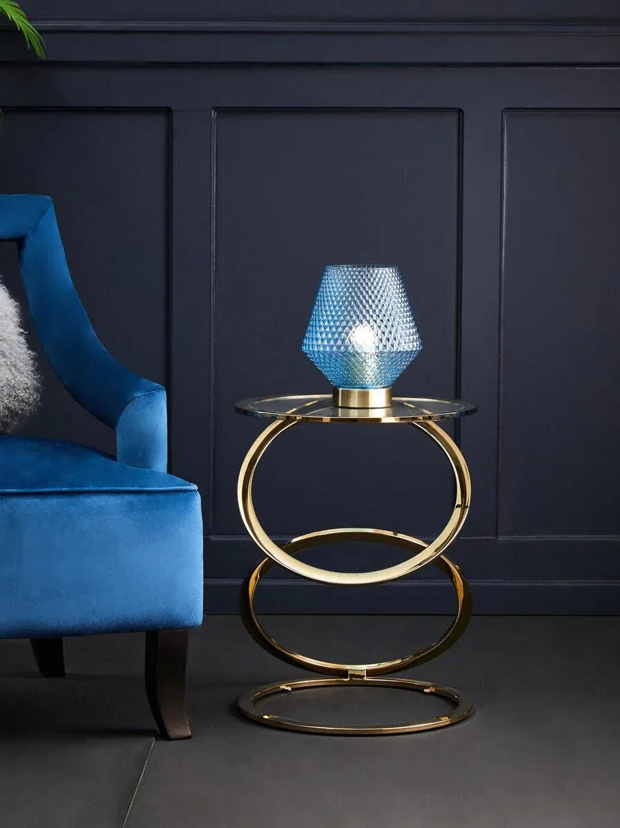 Gold metal side table with glass top beside blue velvet chair and blue textured table lamp