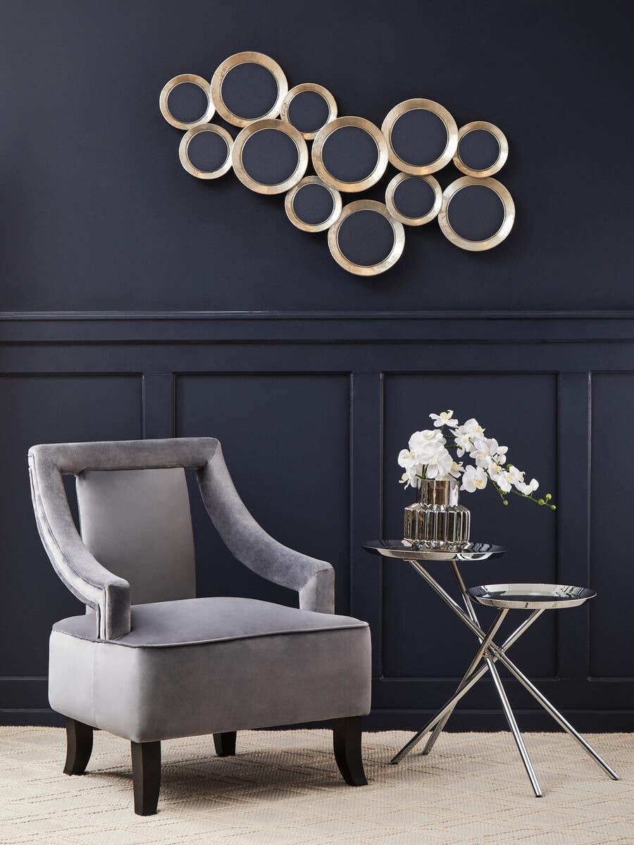 Grey velvet accent chair with black legs beside chrome nesting side tables and wall art