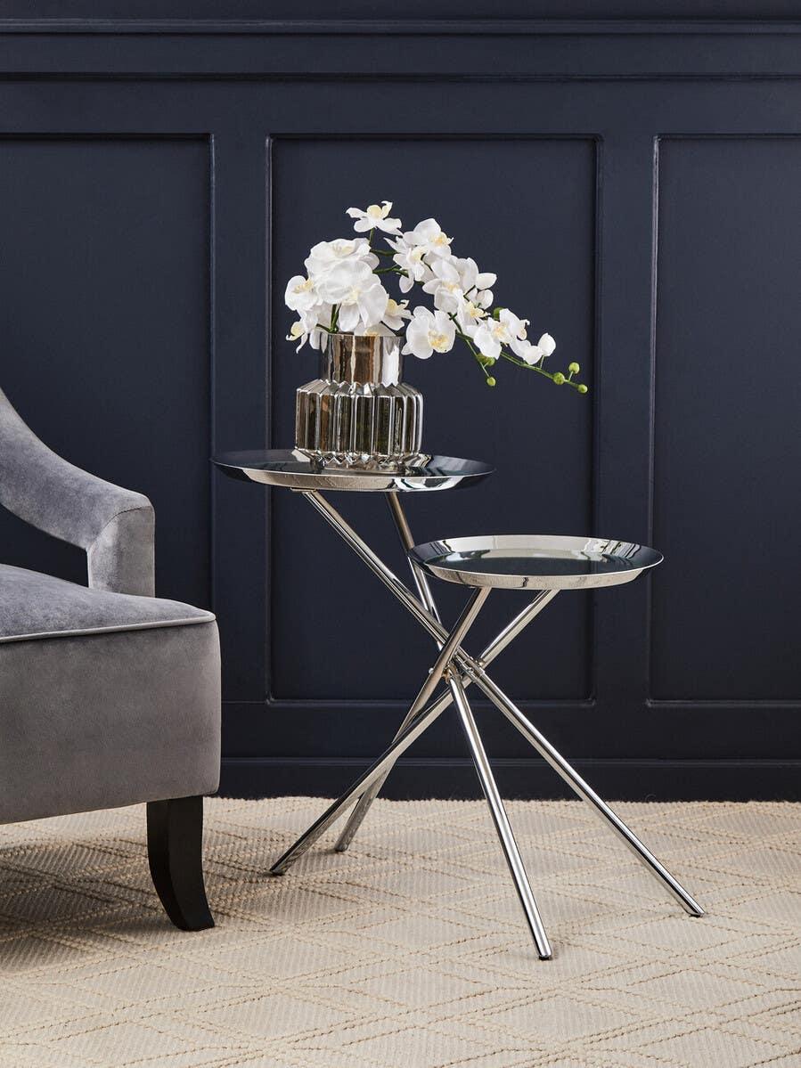 Pair of chrome side tables with glass vase of white flowers beside grey velvet armchair