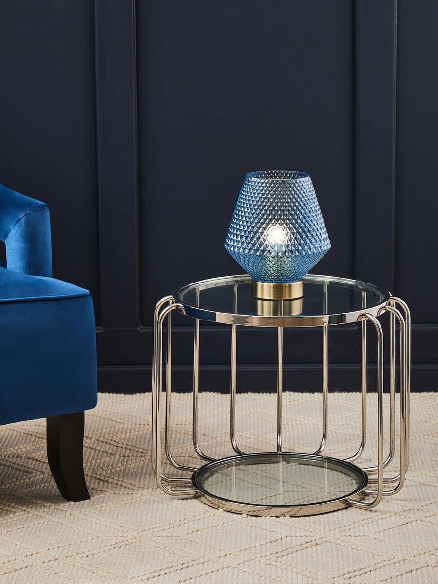 Glass side table with metal frame and textured blue table lamp beside blue velvet chair