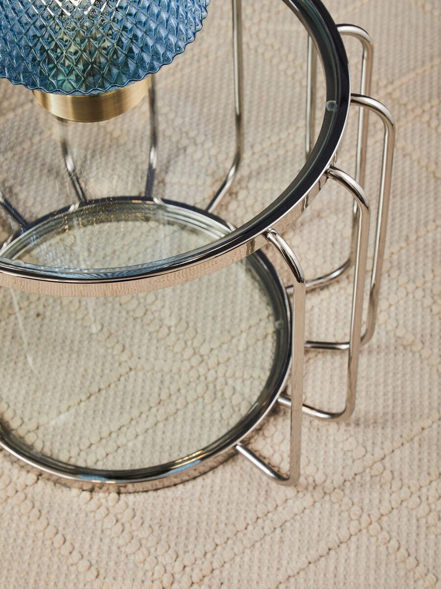Round glass side table with chrome legs on textured cream rug, blue lamp on top