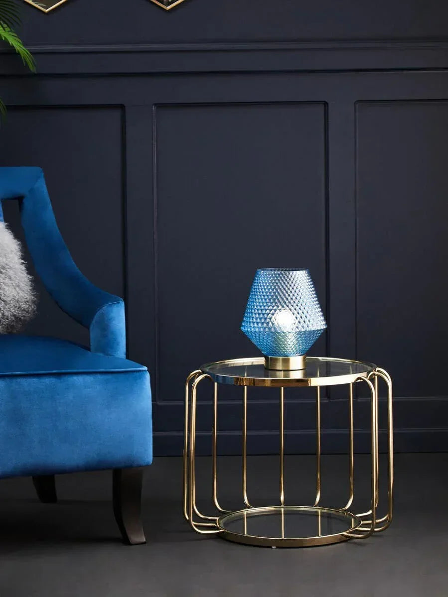 Glass top side table with gold metal frame and blue table lamp beside blue velvet armchair
