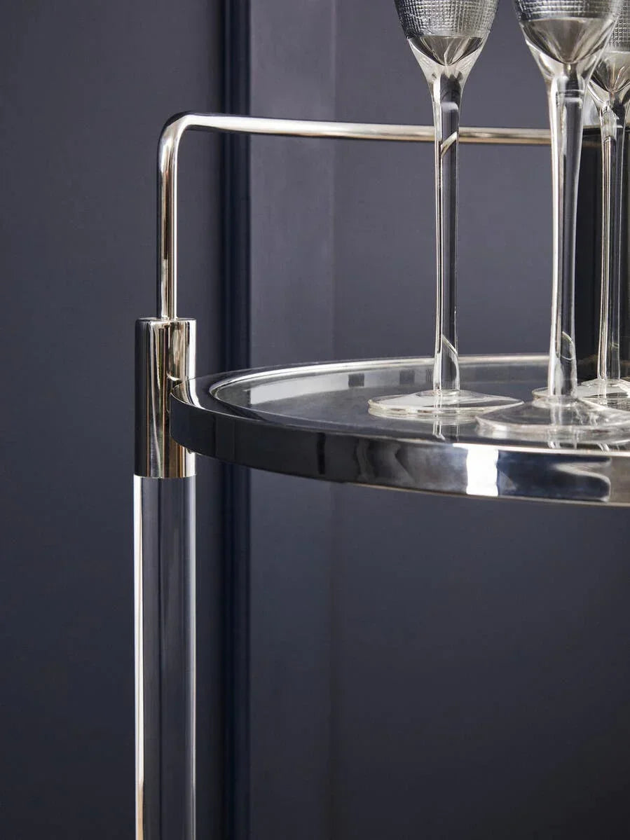 Chrome and glass bar cart with tall wine glasses on circular shelf against dark wall