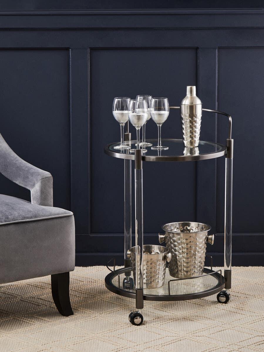Glass and metal drinks trolley with wine glasses, shaker, and ice buckets beside a grey armchair