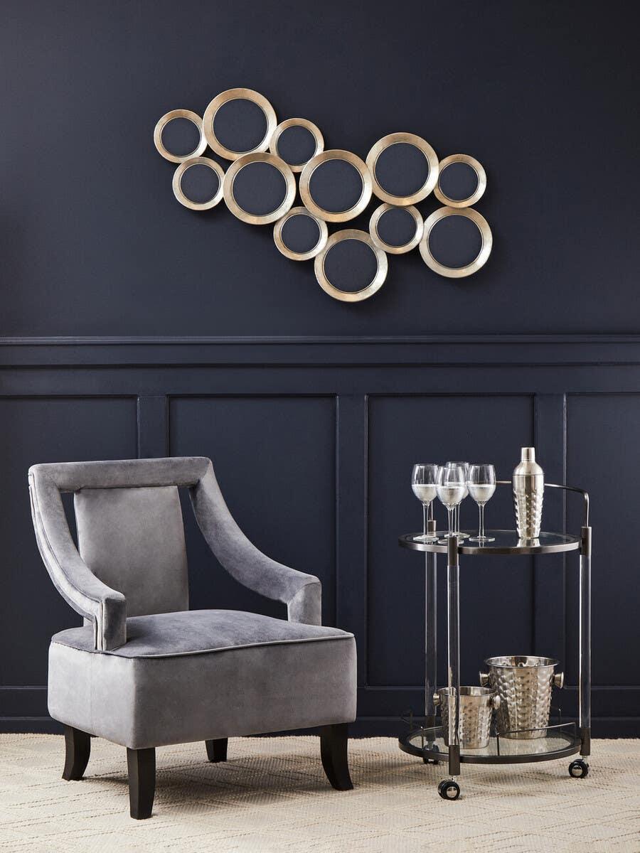 Grey velvet accent chair and metal bar cart with glassware against navy wall with gold wall art