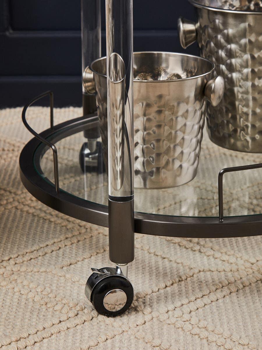 Glass bar cart with metal bucket on textured rug, chrome frame and caster wheel detail