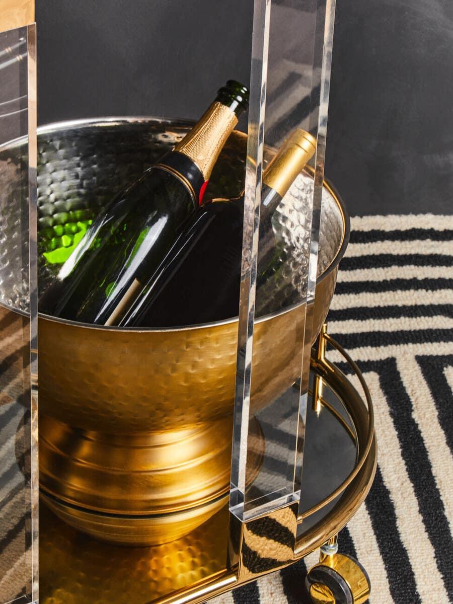 Gold hammered metal wine cooler with bottles on brass bar cart, patterned rug underneath