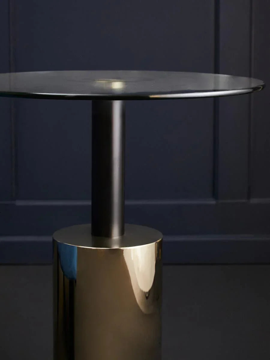 Round side table with black glass top and gold cylindrical metal base against dark wall