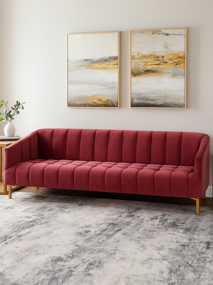 A plush, wine-colored velvet sofa with a modern design, featuring a tufted backrest and wooden legs, positioned in a bright room with two abstract art pieces on the wall and a light-colored area rug beneath it