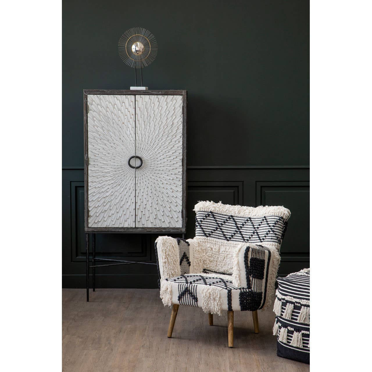 A black and white shaggy pouffe placed in a stylish living room setting, featuring a dark green wall, a light-colored wooden floor, and a patterned armchair nearby. A tall cabinet with a circular handle is visible in the background, along with a decorative light fixture above