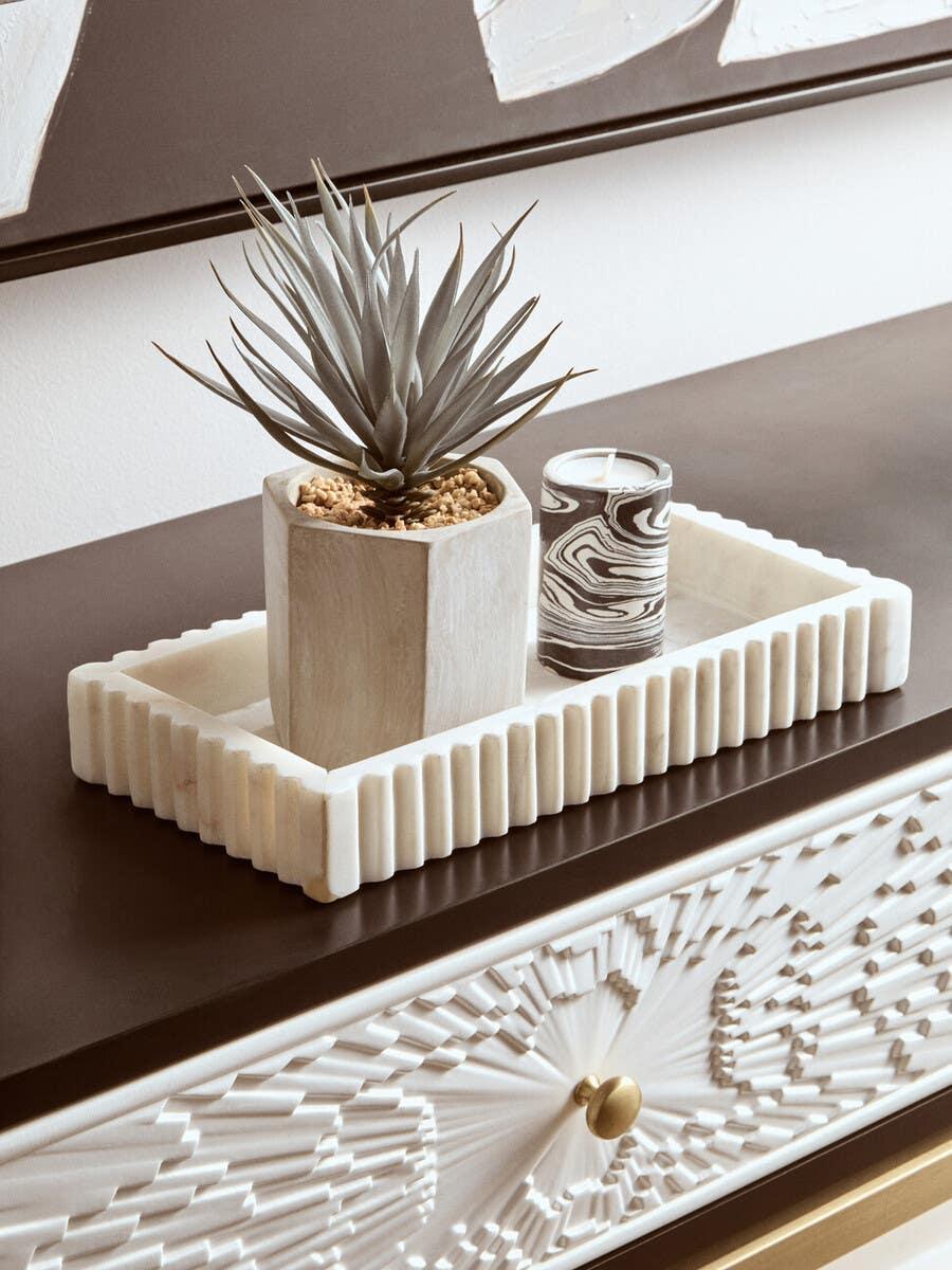White ceramic tray with potted artificial plant and patterned candle on carved console table