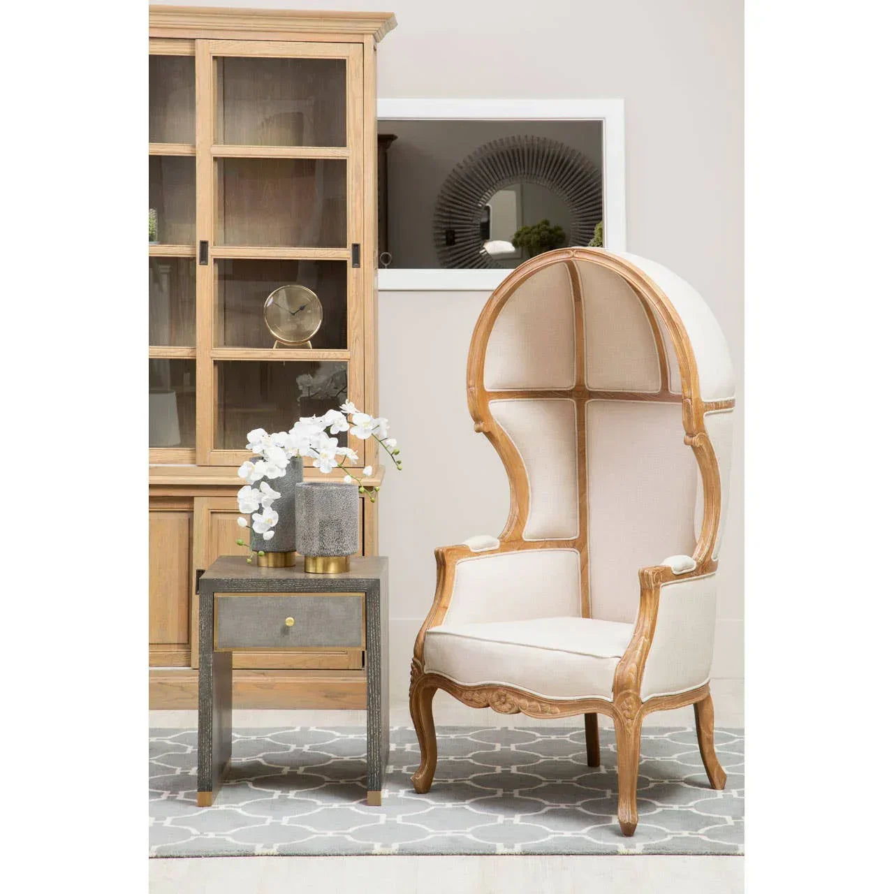 High back canopy accent chair with light wood frame next to side table and glass cabinet