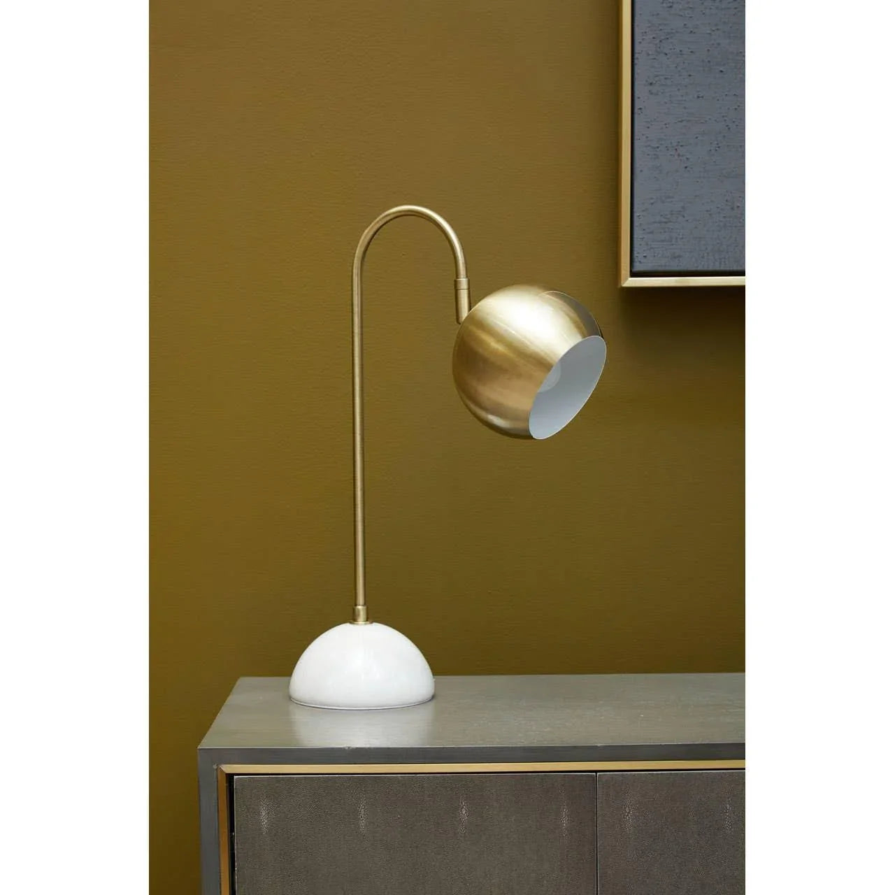 Gold metal desk lamp with curved arm and white base on grey sideboard