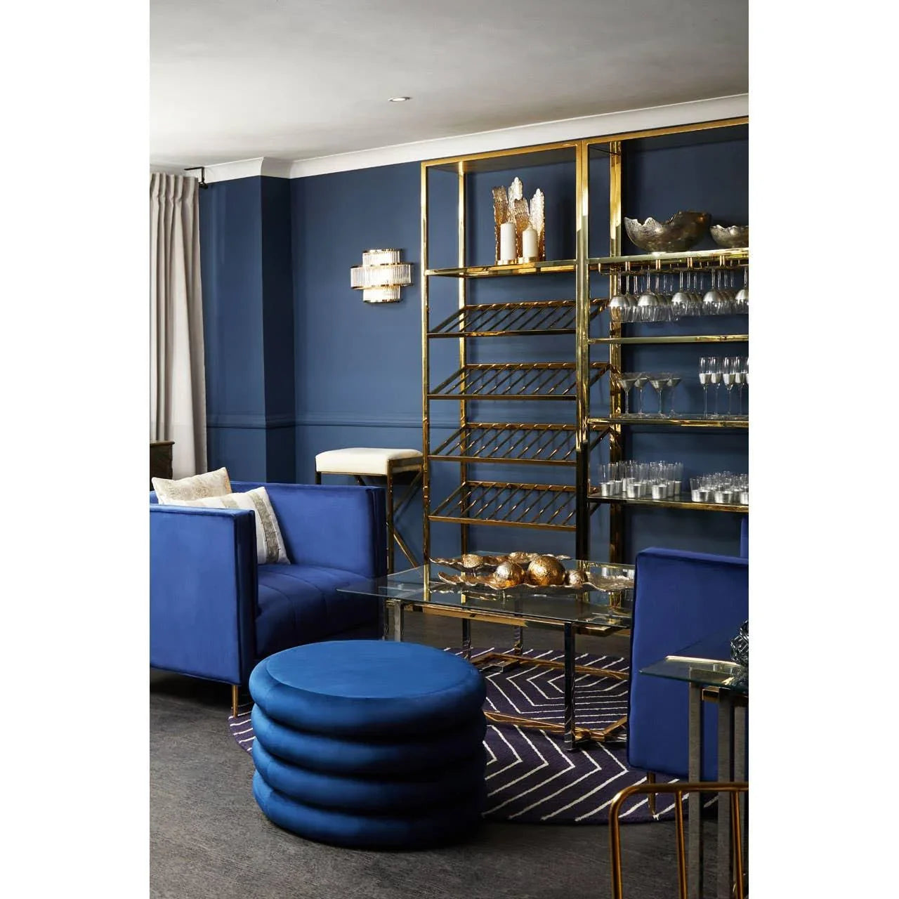 Blue velvet sofa with matching round ottoman, gold shelving, glass coffee table in modern lounge