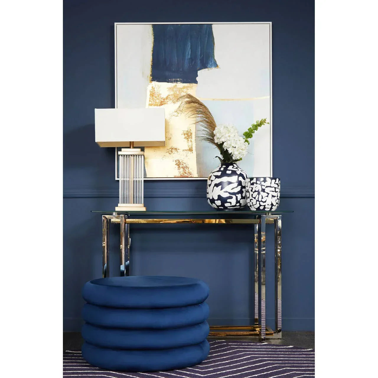 Blue velvet ottoman and glass console table with vases and lamp against navy wall