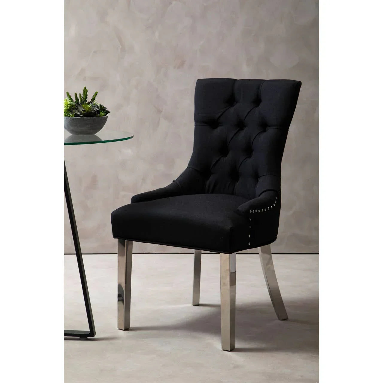 Black upholstered dining chair with button-tufted back and chrome legs beside glass table
