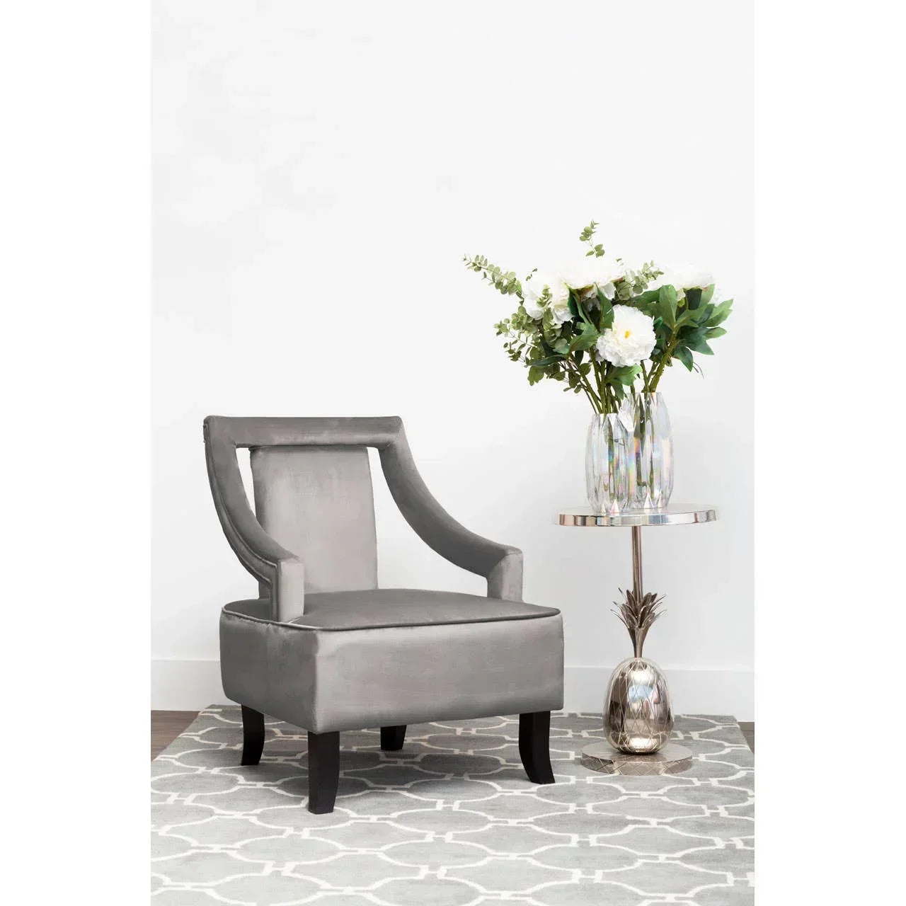 Grey velvet accent chair with curved arms on patterned rug beside metallic side table and flowers