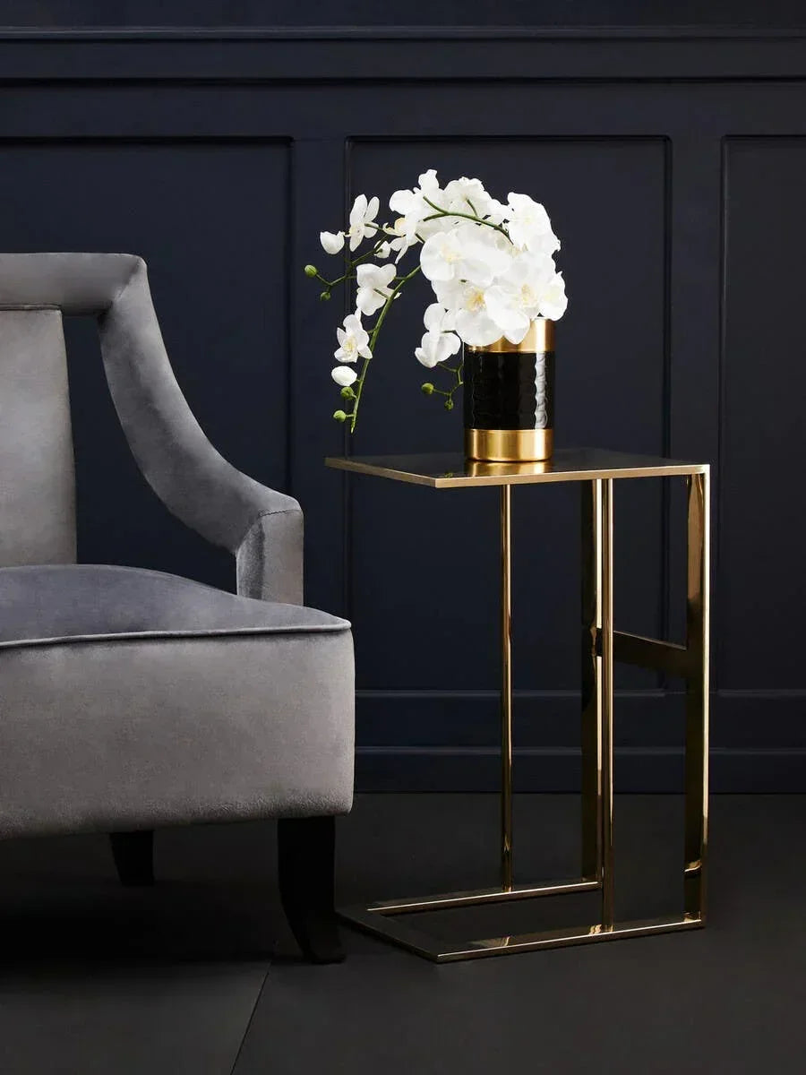 Gold metal side table with black and gold vase holding white orchids beside grey velvet sofa
