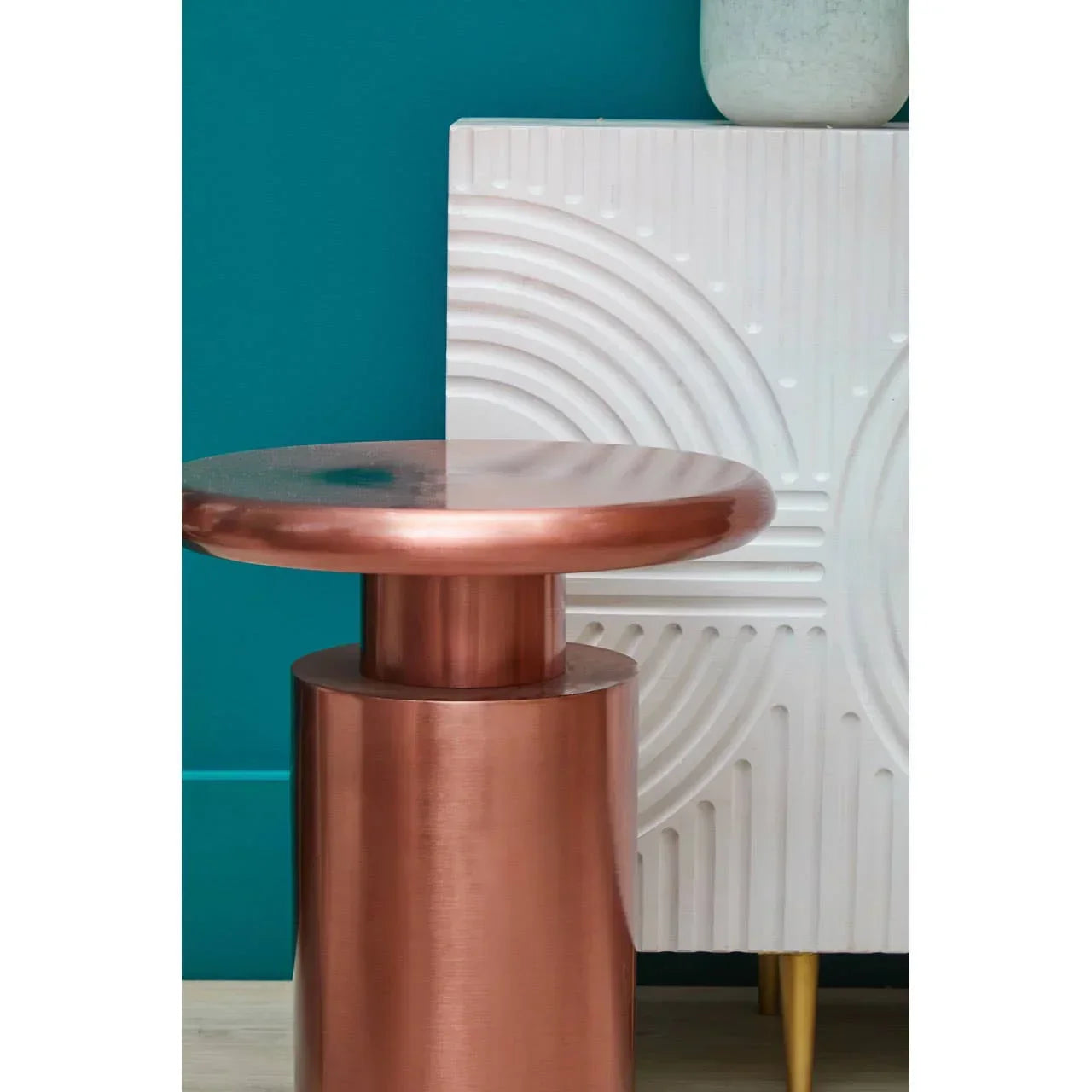 Copper metal side table with round top beside white textured cabinet