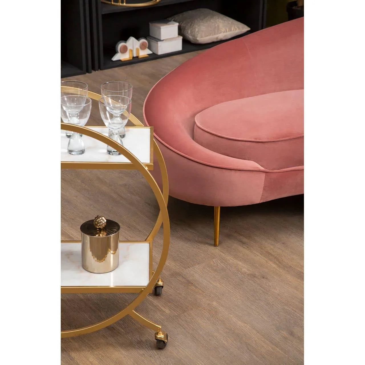 Pink velvet sofa with curved back next to gold drinks trolley with glassware on wooden floor