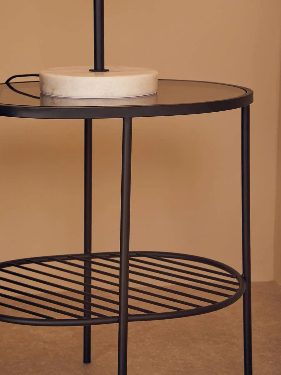 Black metal side table with glass top and slatted lower shelf against neutral wall