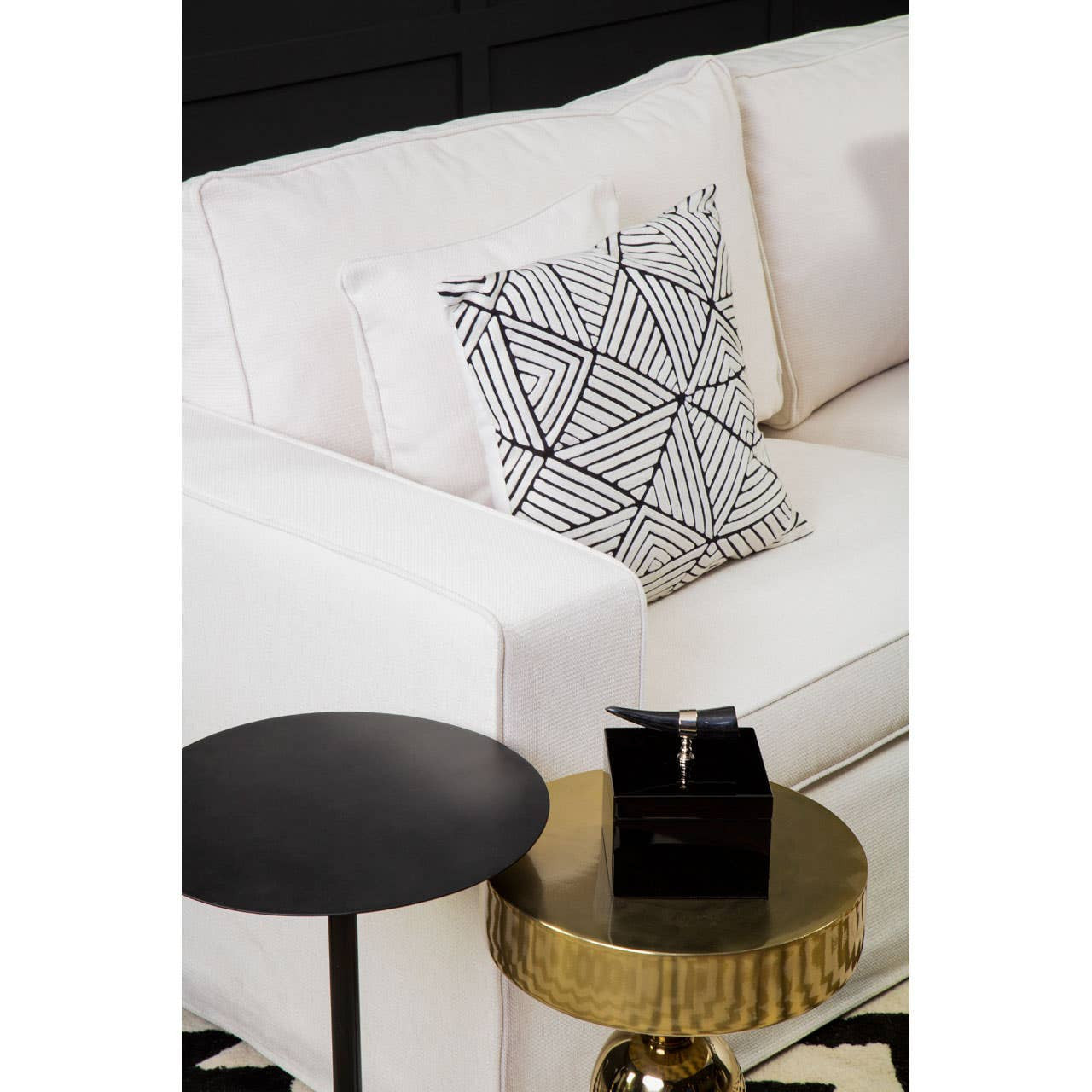A cream fabric sofa with three seats, featuring a decorative geometric patterned pillow, is positioned next to a small black side table and a round gold coffee table with a stack of black books on top. The background is dark, enhancing the sofa's light color