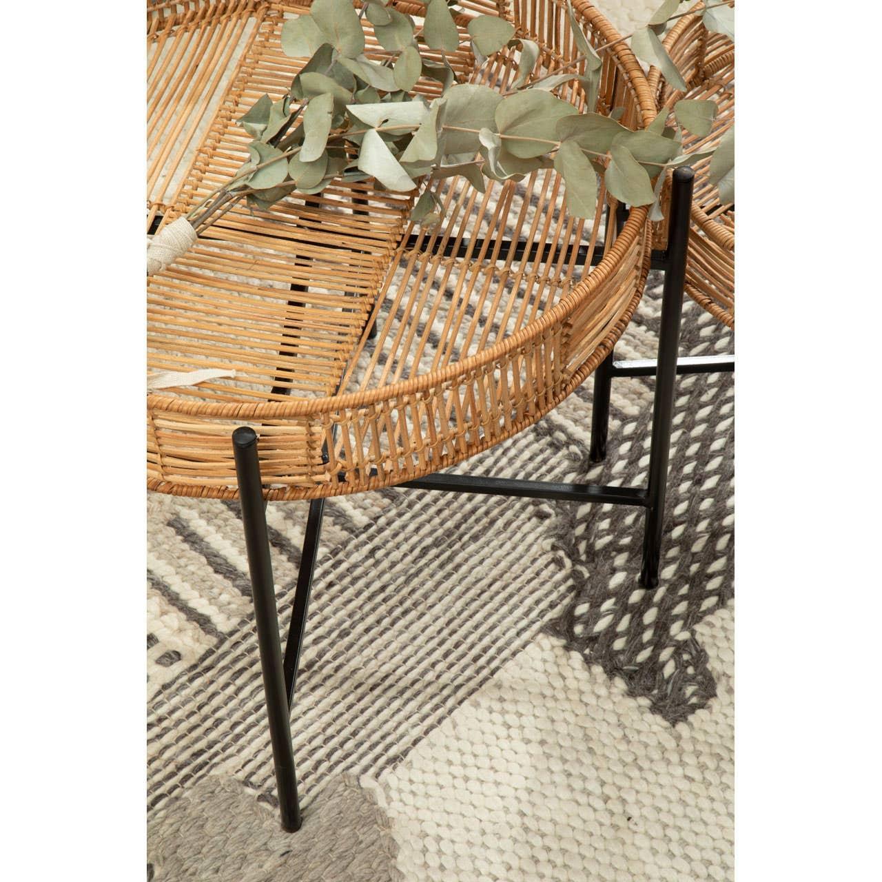 Round rattan side table with black metal legs on patterned rug, decorative leaves on top