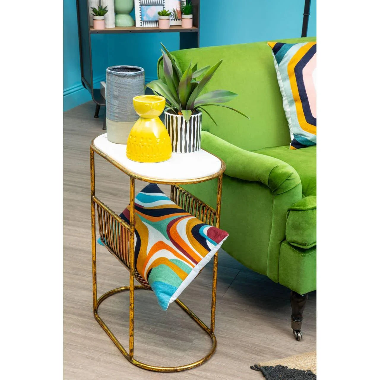 Gold metal side table with white top and shelf beside green velvet sofa and geometric cushions