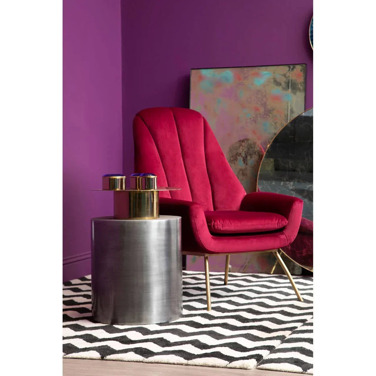 Pink velvet armchair with gold legs beside round metallic accent table on chevron rug