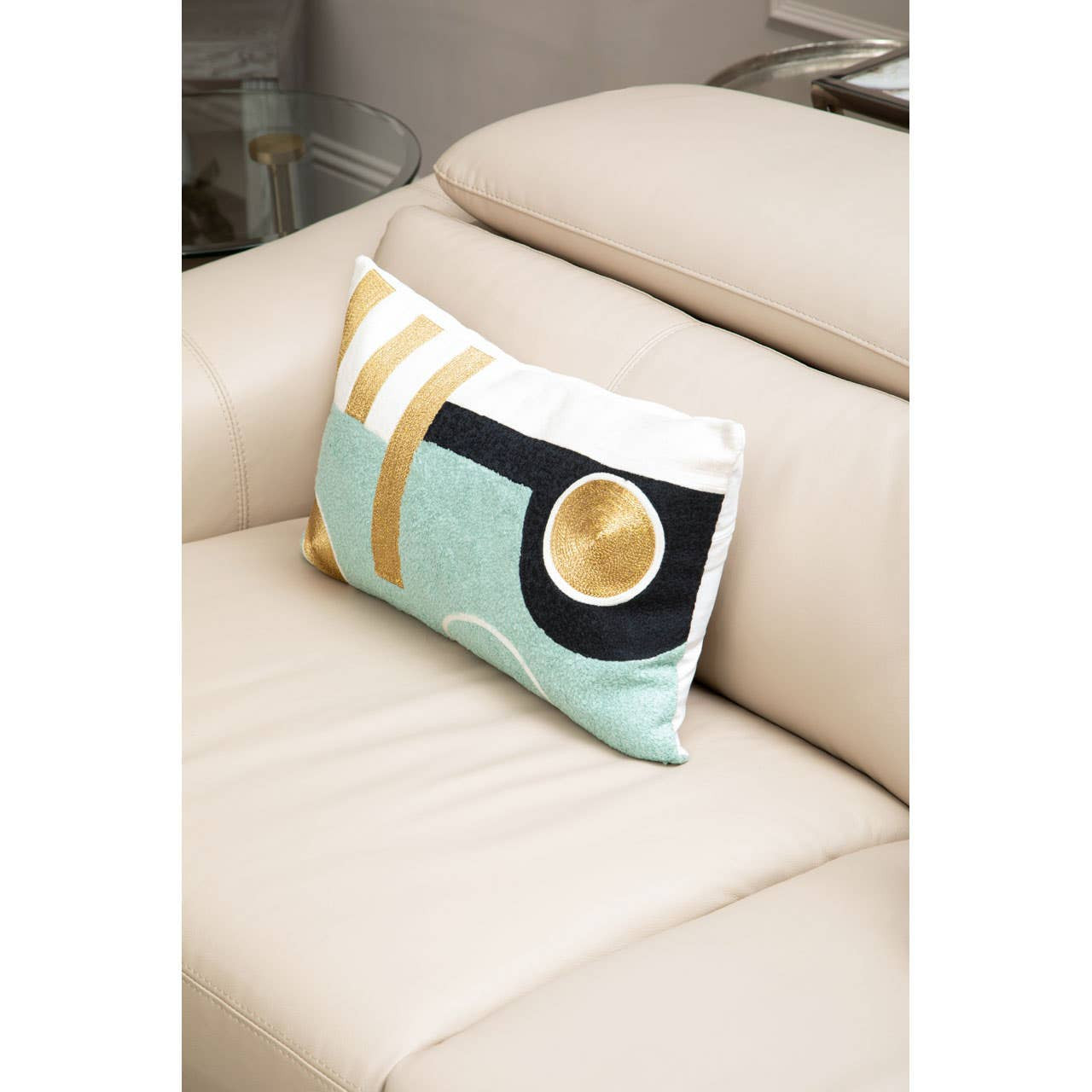 A decorative pillow with a geometric design in mint green, black, and gold rests on the arm of a light stone leather sofa. The sofa features sleek lines and adjustable headrests, showcasing a modern and stylish appearance