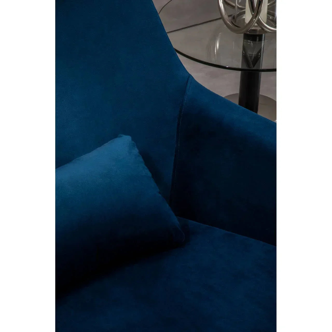 Blue velvet armchair with matching cushion beside round glass coffee table