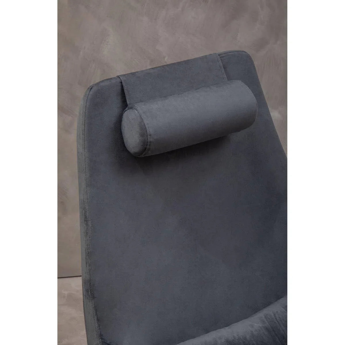Grey velvet chair with curved backrest and cylindrical headrest against neutral wall