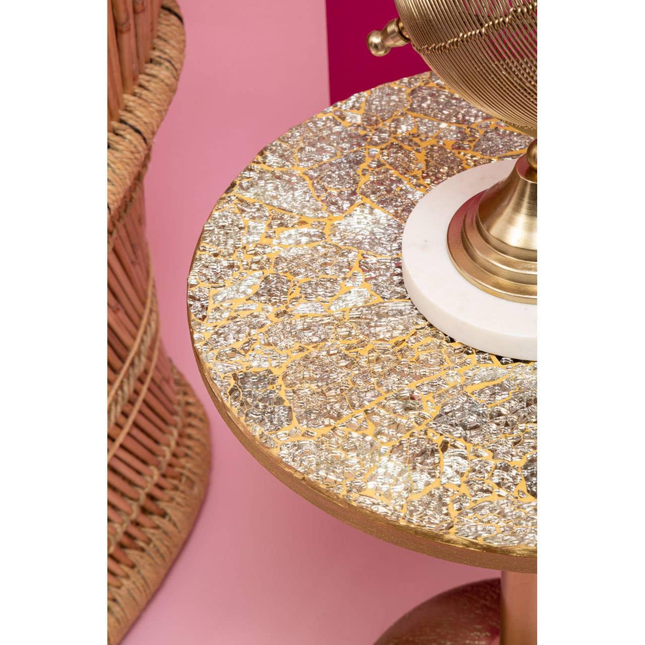 Round side table with gold and silver mosaic top next to wicker chair on pink background