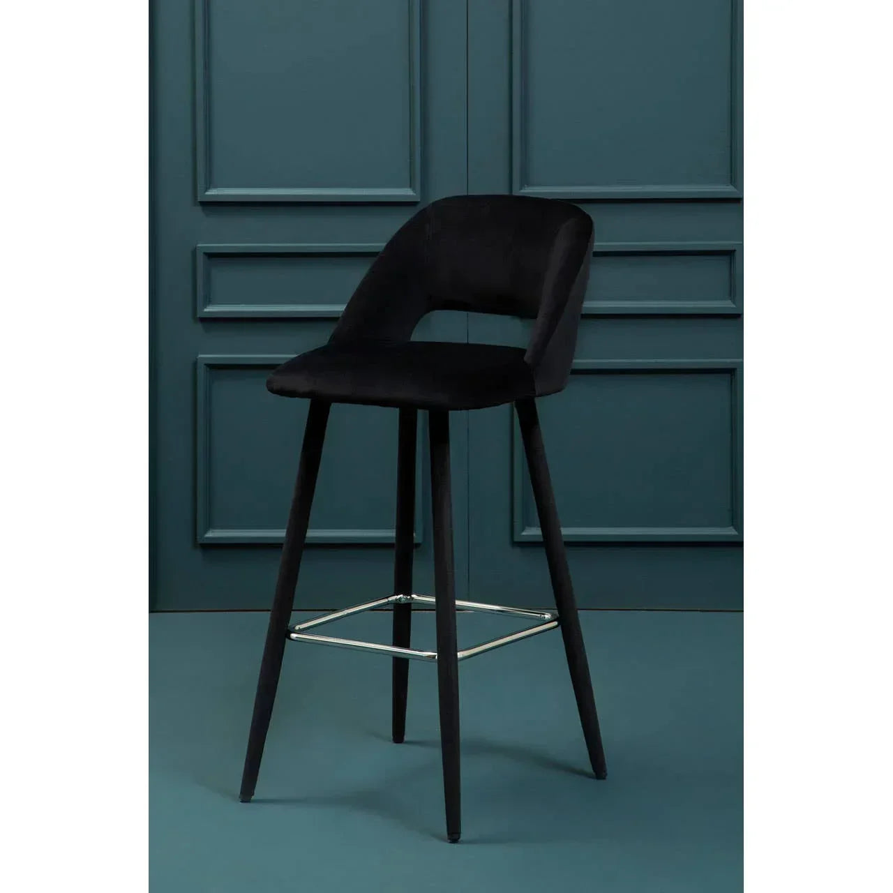 Black velvet bar stool with curved backrest and metal footrest against blue panel wall