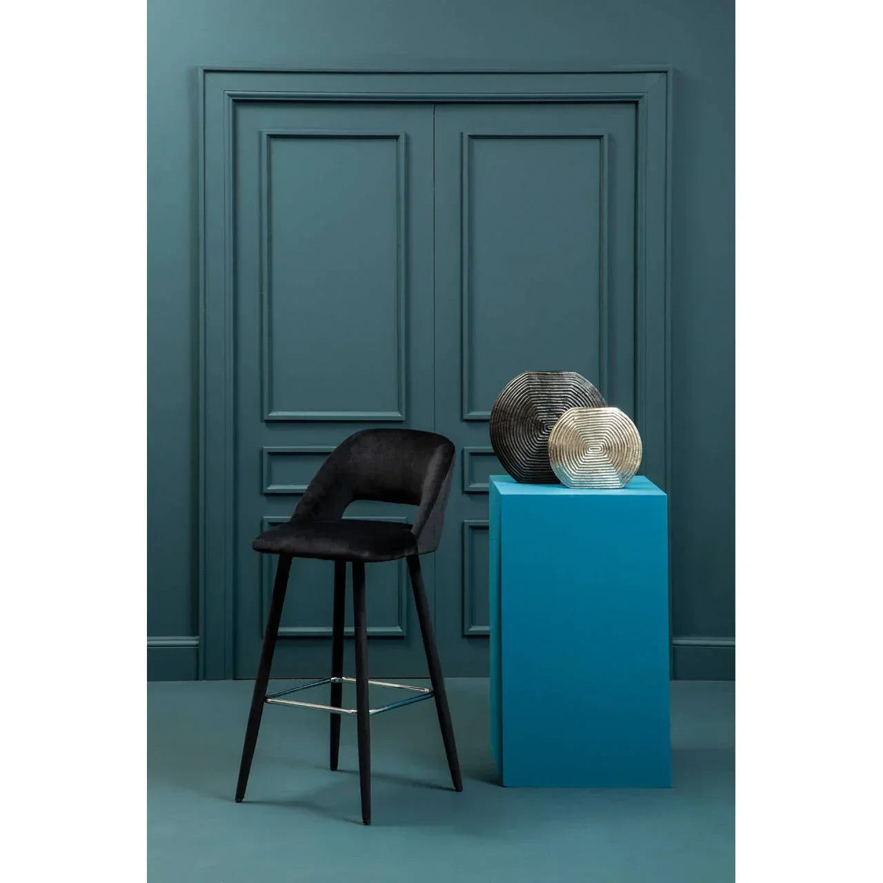 Black velvet bar stool with curved backrest beside modern vases on a blue pedestal