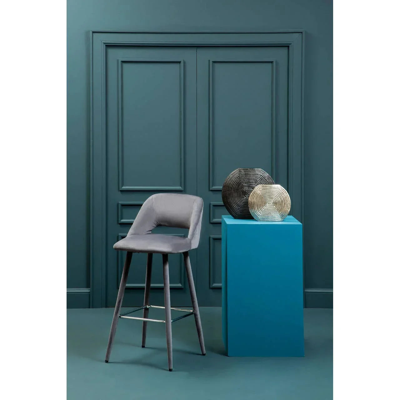 Grey velvet bar stool with metal footrest beside blue pedestal and decorative vases