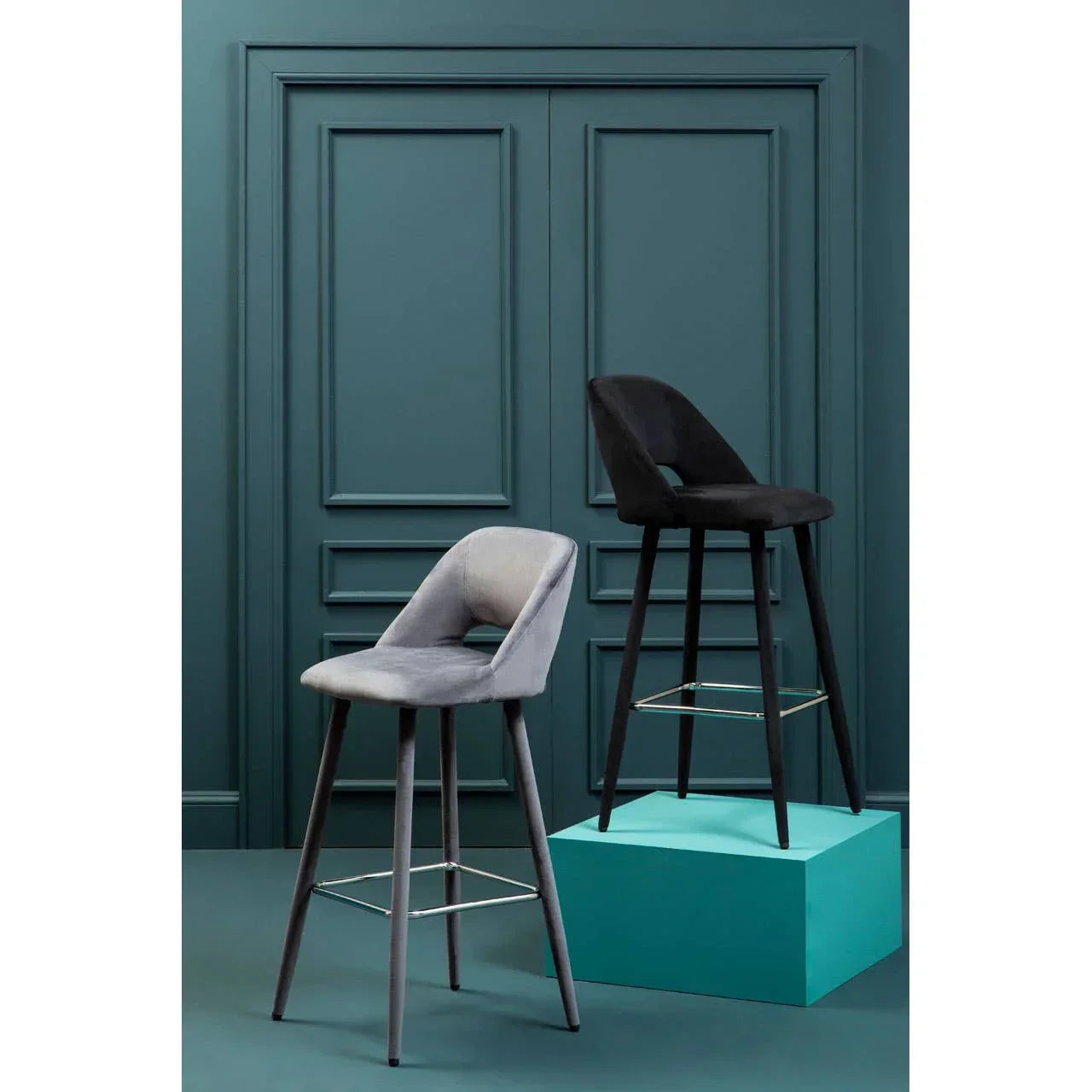 Grey and black upholstered bar stools with metal footrests, set against teal paneled wall