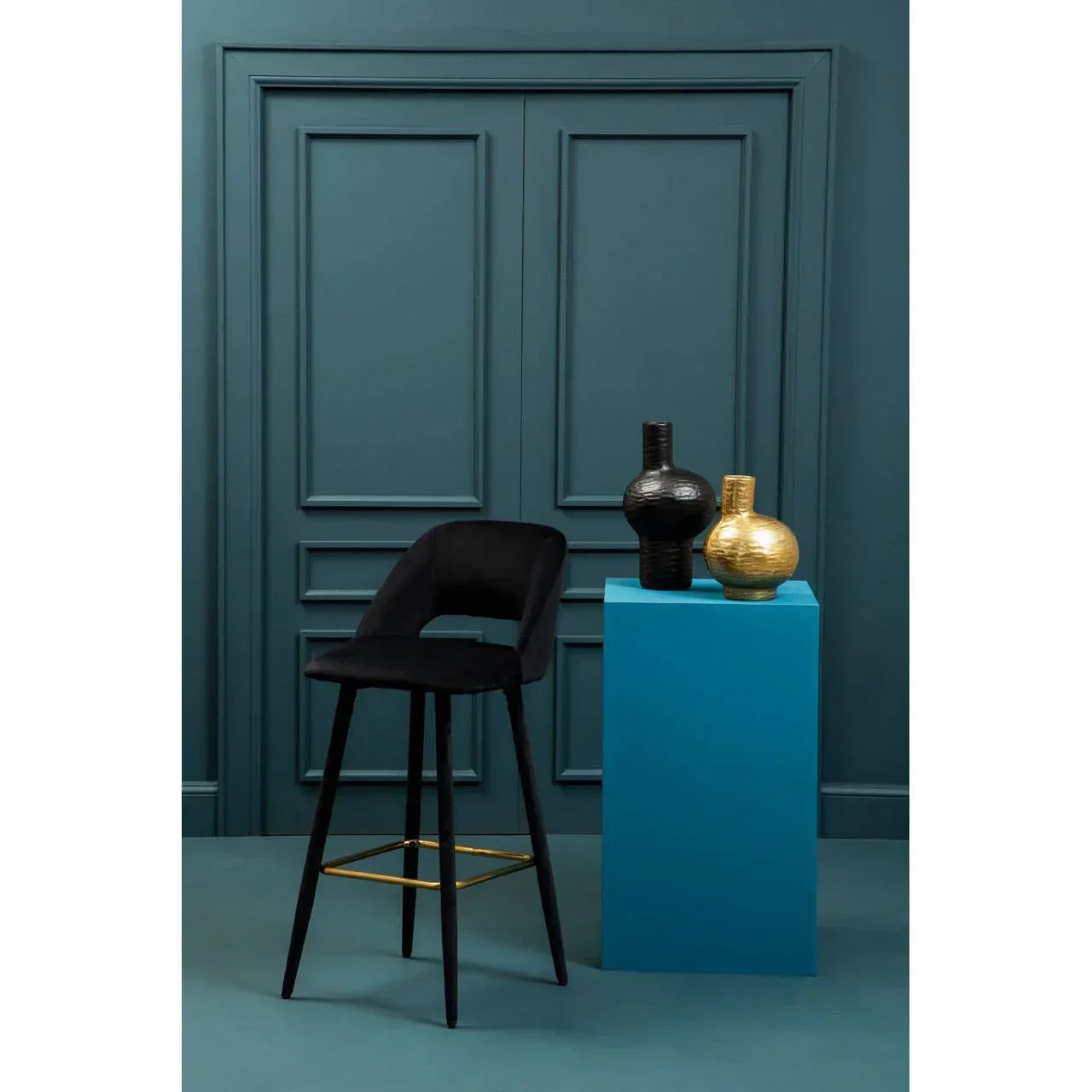 Black velvet bar stool with gold footrest beside blue pedestal displaying black and gold vases