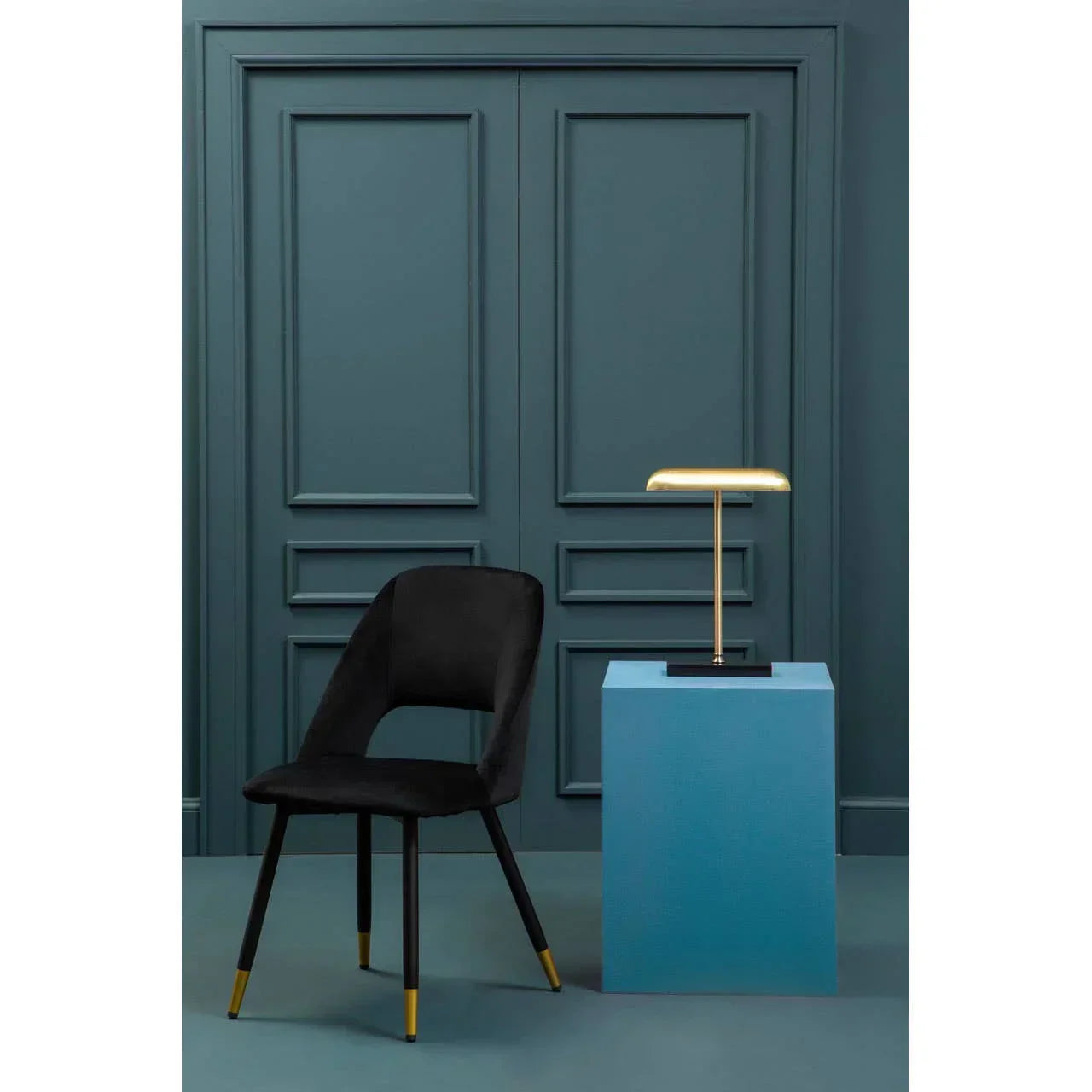 Black velvet dining chair with gold tip legs beside a brass table lamp on blue pedestal